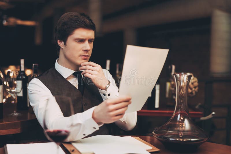 Młody sommelier patrzeje zamyślenie przy wino listy obsiadaniem przy stołem z wino dekantatorem zdjęcia royalty free Młody sommelier patrzeje zamyślenie przy wino listy obsiadaniem przy stołem z wino dekantatorem zdjęcia royalty free