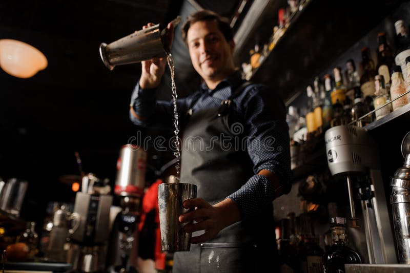 Młody uśmiechnięty barman nalewa alkoholicznego napój od jeden metalu szkła w inny obrazy stock Młody uśmiechnięty barman nalewa alkoholicznego napój od jeden metalu szkła w inny obrazy stock