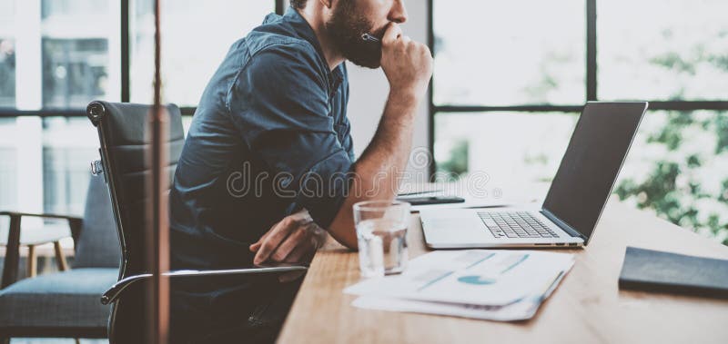 Młody zadumany coworker pracuje przy pogodnym miejsca pracy loft podczas gdy siedzący przy drewnianym stołem Mężczyzna analizuje zdjęcie stock Młody zadumany coworker pracuje przy pogodnym miejsca pracy loft podczas gdy siedzący przy drewnianym stołem Mężczyzna analizuje zdjęcie stock