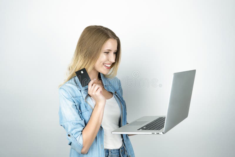 Młodych blond kobiet spojrzeń szczęśliwy mienie jej bank karta w jeden laptopie w inny i ręce, online zakupy, odosobniony biel fotografia stock Młodych blond kobiet spojrzeń szczęśliwy mienie jej bank karta w jeden laptopie w inny i ręce, online zakupy, odosobniony biel fotografia stock