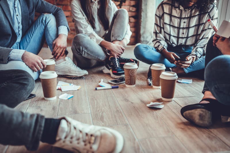 młodzi przedsiębiorcy w ubraniach dorywczych dyskutują o pracy, piją kawę i uśmiechają się siedząc na podłodze w biurze