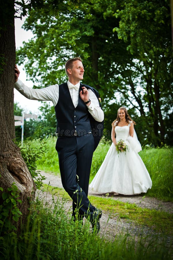 Handsome groom with his blurred pretty dark and long haired bride is leaning at a big tree leaf and looking into the sky. The newlyweds are very cute and pretty. She is wearing a nice white and long wedding dress with nice bouquet and flowers. Handsome groom with his blurred pretty dark and long haired bride is leaning at a big tree leaf and looking into the sky. The newlyweds are very cute and pretty. She is wearing a nice white and long wedding dress with nice bouquet and flowers