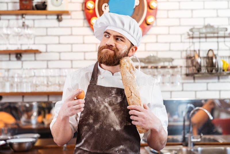 Nachdenklicher bärtiger Bäcker, der Eier und Brot auf der Küche hält stockfoto Nachdenklicher bärtiger Bäcker, der Eier und Brot auf der Küche hält stockfoto