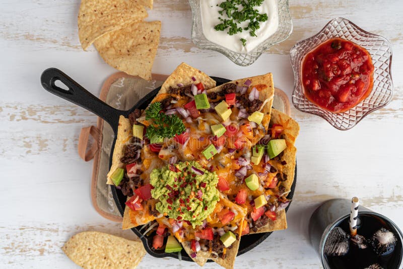 Corn nachos baked in the oven and topped witn ground beef, vegetables, cheddar cheese, and guacamole. Crispy snack with salsa and sour cream dips and coke. Nachos in cast iron skillet on white rustic wooden background. Mexican food. Close up, top view. Corn nachos baked in the oven and topped witn ground beef, vegetables, cheddar cheese, and guacamole. Crispy snack with salsa and sour cream dips and coke. Nachos in cast iron skillet on white rustic wooden background. Mexican food. Close up, top view.