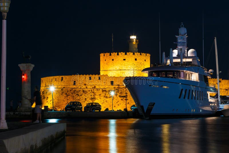 Nachtelijke Marina-haven Mandraki Rhodos-eiland met een groot jacht op de pier op de achtergrond van de oude verrijking, het midd stock afbeelding Nachtelijke Marina-haven Mandraki Rhodos-eiland met een groot jacht op de pier op de achtergrond van de oude verrijking, het midd stock afbeelding