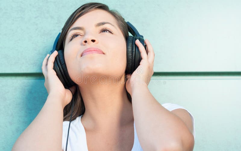 Close-up of young beautiful girl enjoying music on headphones looking up on green wall background with copy text space. Close-up of young beautiful girl enjoying music on headphones looking up on green wall background with copy text space