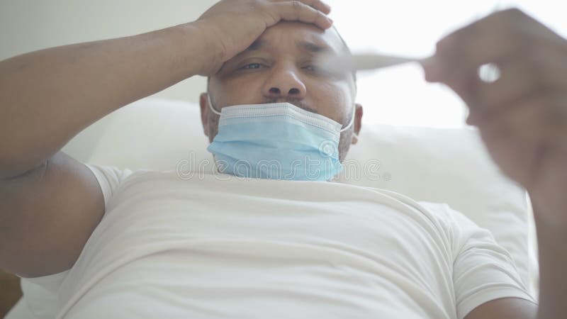 Nahaufnahme des kranken Afroamerikanermannes in der Gesichtsmaske mit Kopfschmerzen und Fieber, das Thermometer betrachtet. Porträ