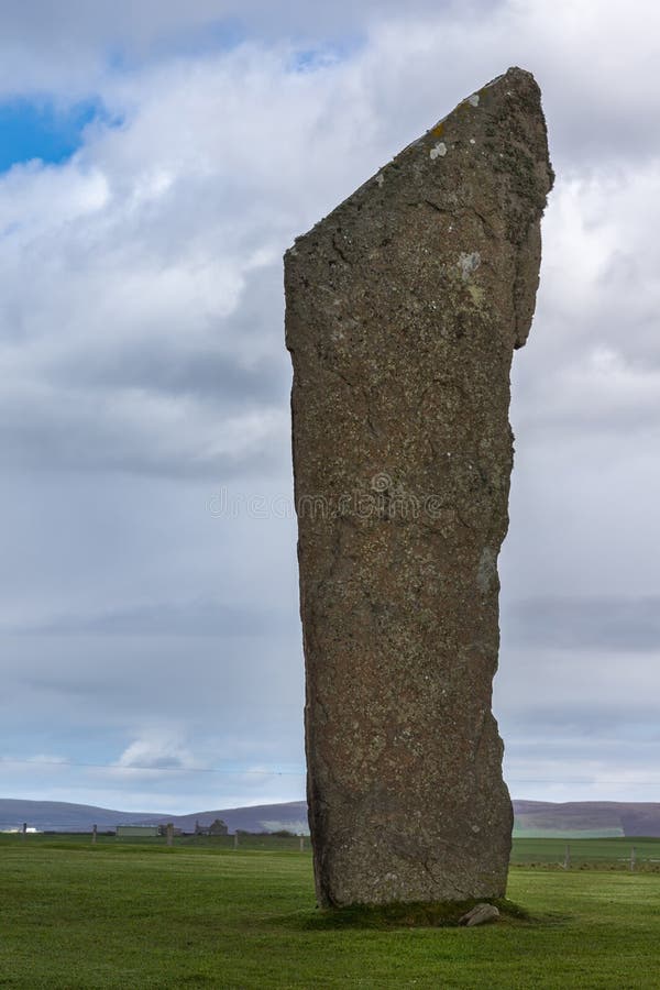 Orkneys, Scotland - June 5, 2012: Ring of Brodgar Neolithic Stone Circle. Closeup of menhir with white and yellow mold spots stands erect on a grass field under blue cloudy sky. Hills and farm in the background. Orkneys, Scotland - June 5, 2012: Ring of Brodgar Neolithic Stone Circle. Closeup of menhir with white and yellow mold spots stands erect on a grass field under blue cloudy sky. Hills and farm in the background.