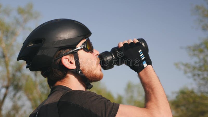 Nahes hohes Porträt von triathlete im schwarzen Sturzhelm und in Trinkwasser der Gläser Bärtiger männlicher Radfahrer trinkt Wass