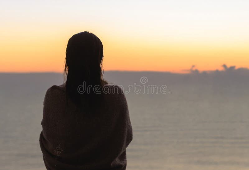 Naked beautiful female with wet hair wrapped in a blanket with cup of coffee standing on the glass balcony early morning at sunris