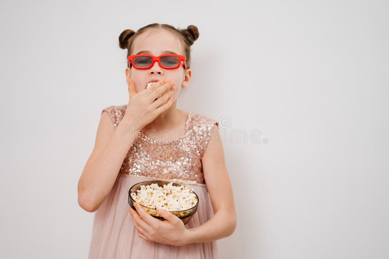 A teen girl with glasses to watch 3D movies eats popcorn stands against a white wall. Home or online cinema. harmful snacking. A teen girl with glasses to watch 3D movies eats popcorn stands against a white wall. Home or online cinema. harmful snacking.