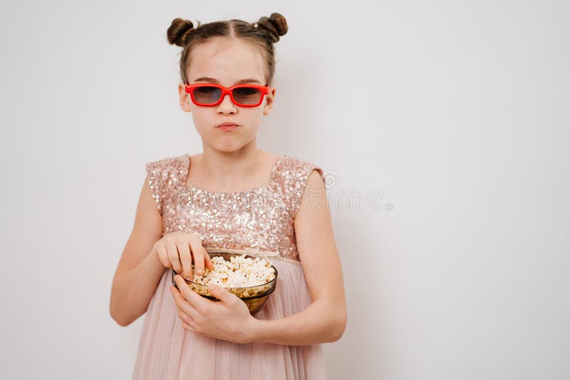 A teen girl with glasses to watch sad 3D movies eats popcorn stands against a white wall. Home or online cinema. harmful snacking. A teen girl with glasses to watch sad 3D movies eats popcorn stands against a white wall. Home or online cinema. harmful snacking.