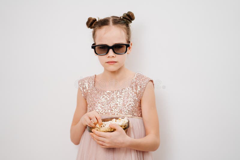 A teen girl with glasses to watch sad 3D movies eats popcorn stands against a white wall. Home or online cinema. harmful snacking. A teen girl with glasses to watch sad 3D movies eats popcorn stands against a white wall. Home or online cinema. harmful snacking.