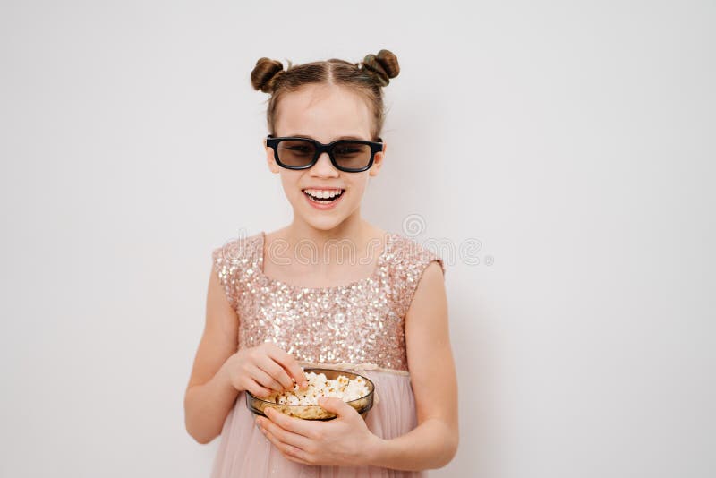 A teen girl with glasses to watch funny 3D movies eats popcorn stands against a white wall. Home or online cinema. harmful snacking. A teen girl with glasses to watch funny 3D movies eats popcorn stands against a white wall. Home or online cinema. harmful snacking.