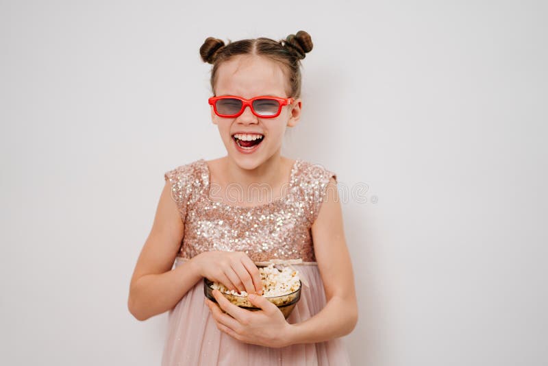 A teen girl with glasses to watch funny 3D movies eats popcorn stands against a white wall. Home or online cinema. harmful snacking. A teen girl with glasses to watch funny 3D movies eats popcorn stands against a white wall. Home or online cinema. harmful snacking.