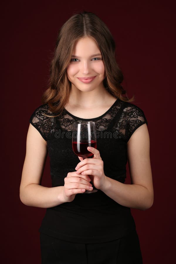 Teen in dress holding glass of wine, girl with wineglass, high fashion look, beautiful girl, brunette girl, isolated, model in studio, long hair, close up, dark red background. Teen in dress holding glass of wine, girl with wineglass, high fashion look, beautiful girl, brunette girl, isolated, model in studio, long hair, close up, dark red background