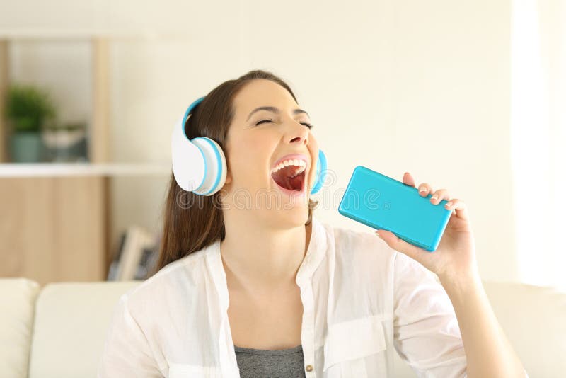 Happy teen singing and listening to music with a smartphone and headphones sitting on a couch in the living room at home. Happy teen singing and listening to music with a smartphone and headphones sitting on a couch in the living room at home