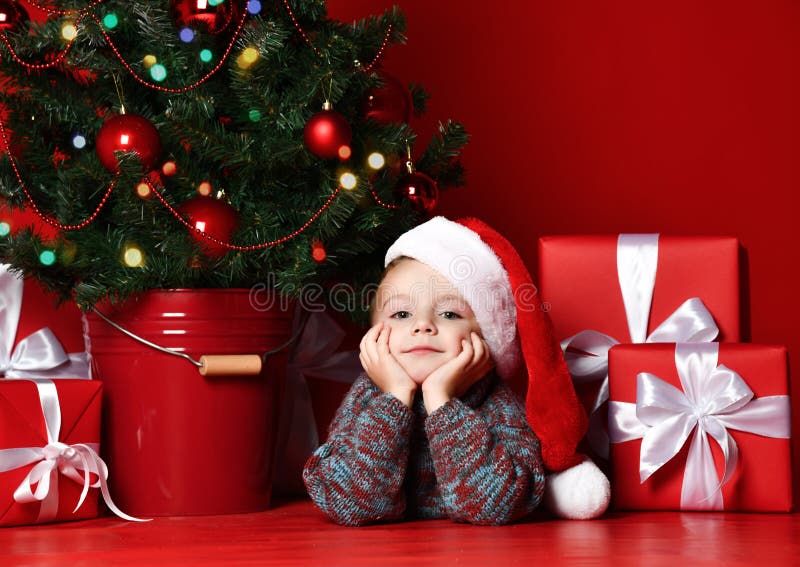 Natale felice e nuovo anno Ritratto del bambino in regali aspettanti di Natale del cappello rosso di Santa immagini stock libere da diritti Natale felice e nuovo anno Ritratto del bambino in regali aspettanti di Natale del cappello rosso di Santa immagini stock libere da diritti