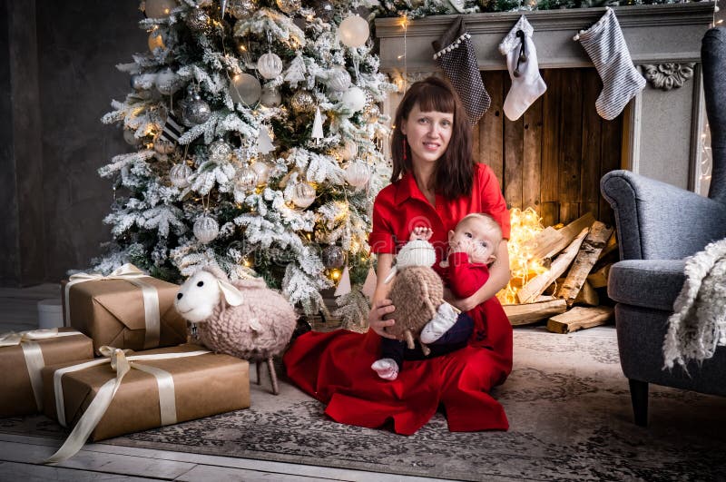 Natale nel circolo famigliare: una giovane madre in un vestito rosso sta sedendosi vicino all'albero di Natale e sta tenendo una fotografia stock Natale nel circolo famigliare: una giovane madre in un vestito rosso sta sedendosi vicino all'albero di Natale e sta tenendo una fotografia stock