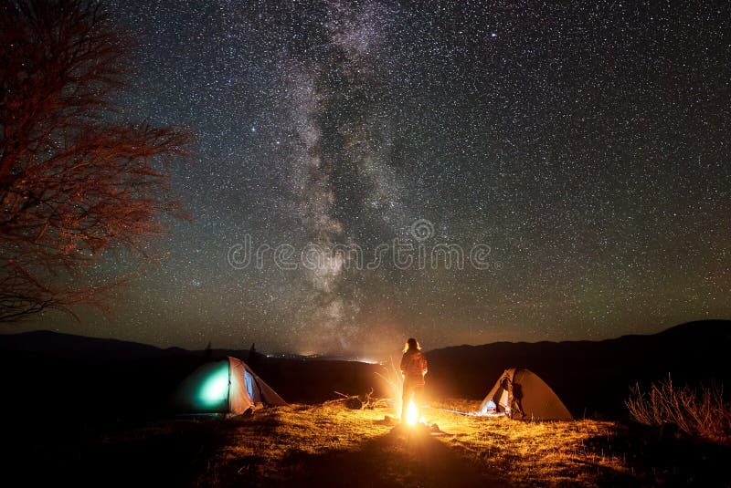 Natt som campar i berg Kvinnlig fotvandrare som vilar nära lägereld, turist- tält under stjärnklar himmel royaltyfri bild Natt som campar i berg Kvinnlig fotvandrare som vilar nära lägereld, turist- tält under stjärnklar himmel royaltyfri bild