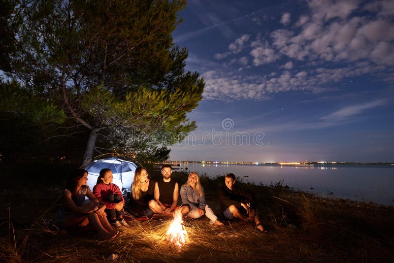 Nattsommar som campar p? kust Grupp av unga turister runt om l?gereld n?ra t?ltet under aftonhimmel royaltyfri bild Nattsommar som campar p? kust Grupp av unga turister runt om l?gereld n?ra t?ltet under aftonhimmel royaltyfri bild