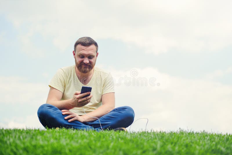 Natur och teknologi En man med ett skägg ligger på ett grönt gräs med en smartphone i hans händer är banken för uppladdningsmakt royaltyfri bild Natur och teknologi En man med ett skägg ligger på ett grönt gräs med en smartphone i hans händer är banken för uppladdningsmakt royaltyfri bild