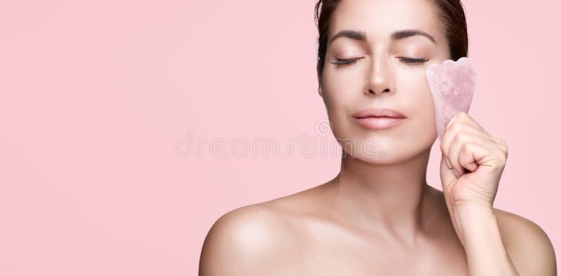 Natural beauty applying gua sha massager on face. Middle aged woman enjoying a beauty face treatment with a quartz pink guasha.