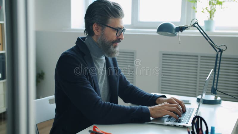 Natuurlijke man in een elegant pak die een bril aantrekt en op kantoor werkt met een laptop