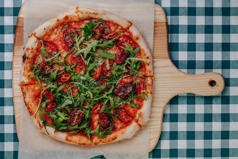 Neapolitan spicy pizza on a cream sauce with ham, cheese, arugula, basil, tomatoes, pepperoni pepper sprayed with cheese on a wooden board on a tablecloth in a cell with a place for text. Neapolitan spicy pizza on a cream sauce with ham, cheese, arugula, basil, tomatoes, pepperoni pepper sprayed with cheese on a wooden board on a tablecloth in a cell with a place for text