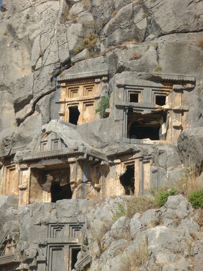 Necropolis of Myra with graves dug in the rock in Turkey. Stones and rocks. Dry vegetation. Grass and shrubs on the rocks. Sunny day. Travel destination. Large number of abodes for the dead ones with niches. Archeological area. Small houses inside the rock. Culture and story. Necropolis of Myra with graves dug in the rock in Turkey. Stones and rocks. Dry vegetation. Grass and shrubs on the rocks. Sunny day. Travel destination. Large number of abodes for the dead ones with niches. Archeological area. Small houses inside the rock. Culture and story.