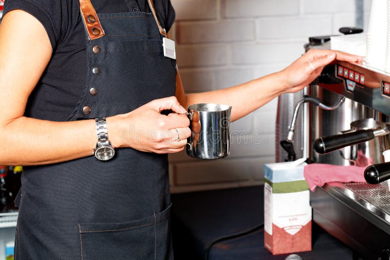 Nelle mani di un barista è una tazza di acciaio inossidabile per la fabbricazione di caffé di latte sullo sfondo di una macchina d. Il barista in un grembiule nero tiene in mano una tazza di metallo per aver fatto caffelatte sullo sfondo di una macchina da caffè e un pacco di crema in sfocatura