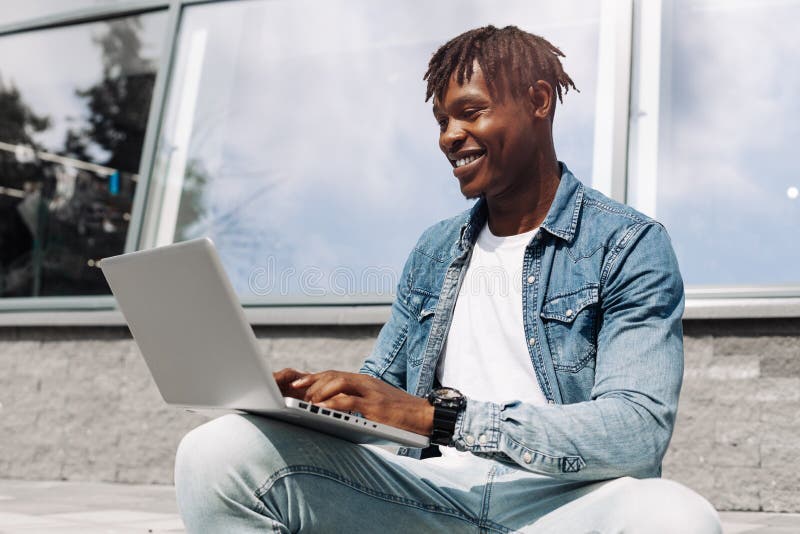 Black African American typing on a laptop on the street in front of a glass building, sitting online, working on the road, high-speed mobile internet, 5g, 4g. Black African American typing on a laptop on the street in front of a glass building, sitting online, working on the road, high-speed mobile internet, 5g, 4g