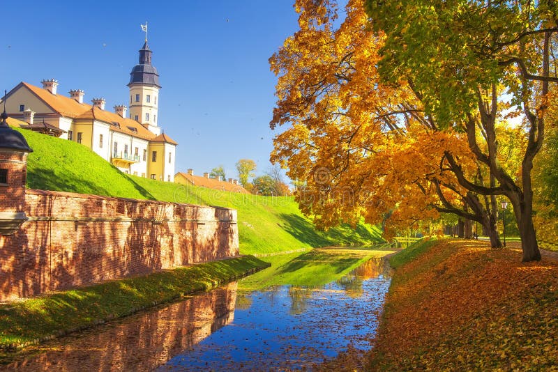 Nesvizh castle at autumn sunny clear day. Colourful yellow and red trees, blue sky, green grass, falling autumnal leaves