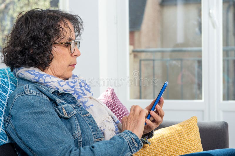 Nette brunette ältere Frau, die Smartphone bei der Sitzung Sofa verwendet stockfotos