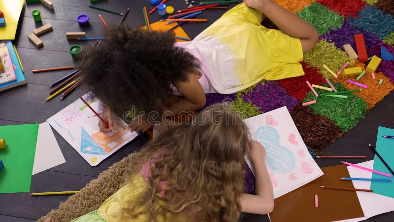 Nette gelockte gemischtrassige Mädchen, die auf Boden liegen und mit Farbbleistiften zeichnen lizenzfreie stockfotos Nette gelockte gemischtrassige Mädchen, die auf Boden liegen und mit Farbbleistiften zeichnen lizenzfreie stockfotos