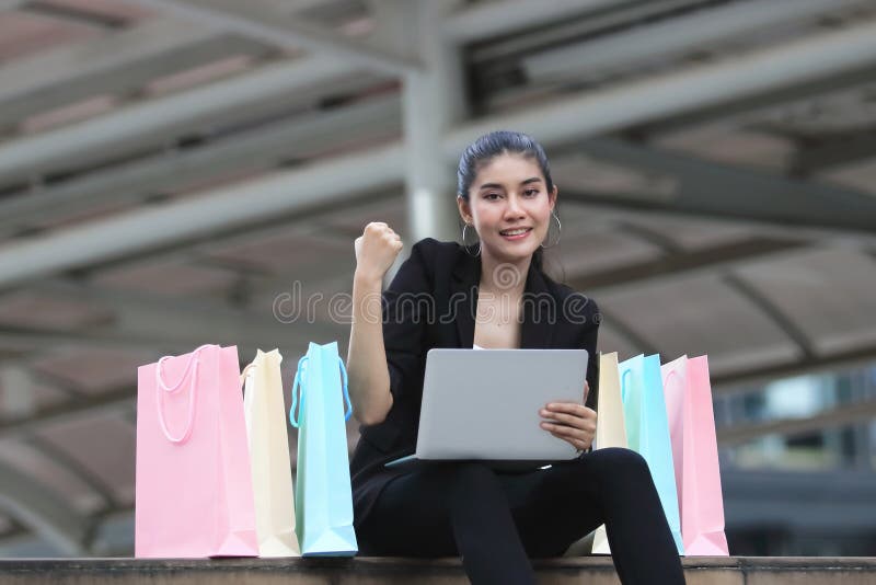 Nette junge Asiatin mit bunter Einkaufstasche und dem Laptop, die an draußen sitzt stockbild Nette junge Asiatin mit bunter Einkaufstasche und dem Laptop, die an draußen sitzt stockbild