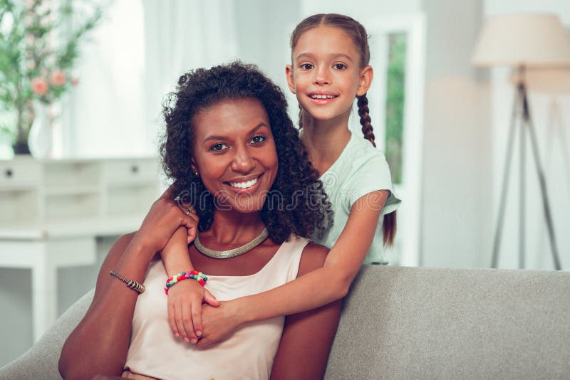 Nette kleine Tochter, die liebevoll afroe-amerikanisch glühende Mutter umarmt lizenzfreies stockfoto