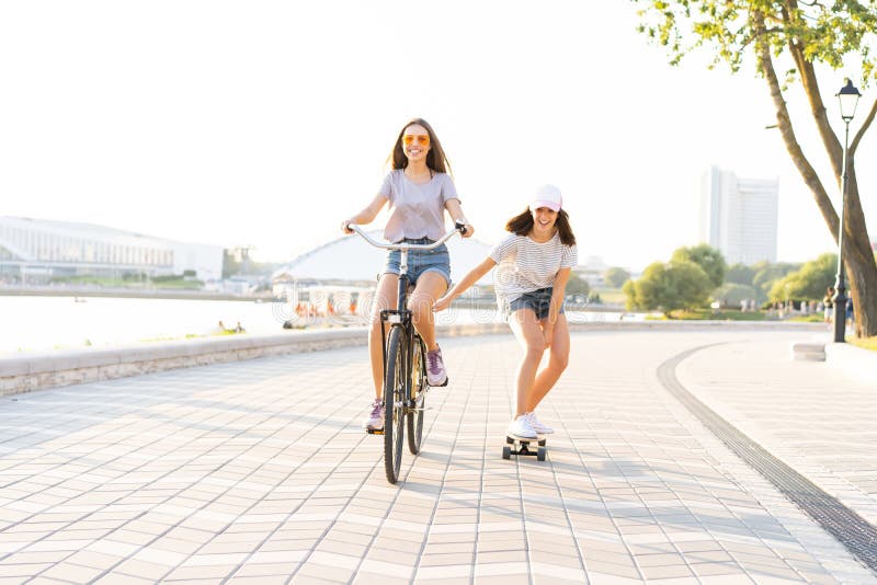 Nette lachende junge Frau in der kurzen Jeanshose auf dem Fahrrad, welches das Mädchen an hält bei der Anwendung des Skateboards