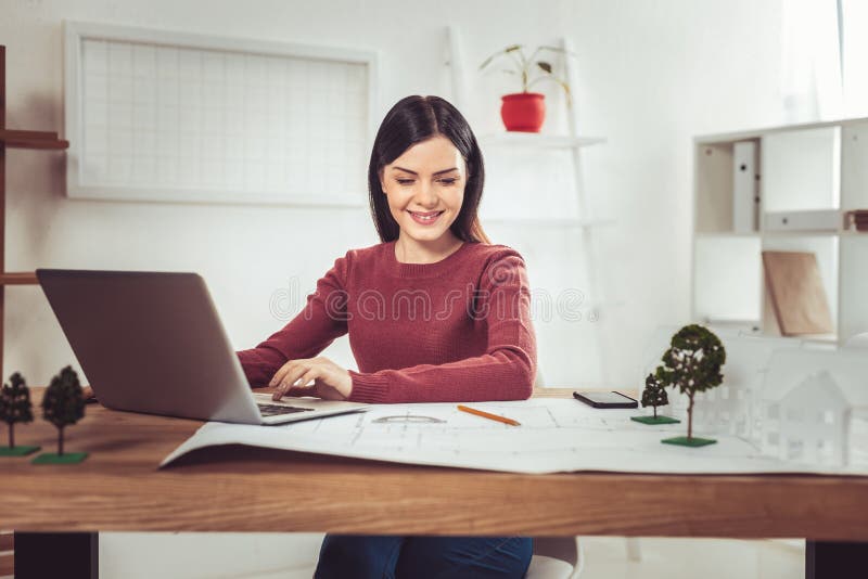 Netter Brunette, der am Computer arbeitet. Lieblingsjob Der junge Lächeln auf ihrem Gesicht haltene und lehnende Architekt bewaffnet auf Tabelle