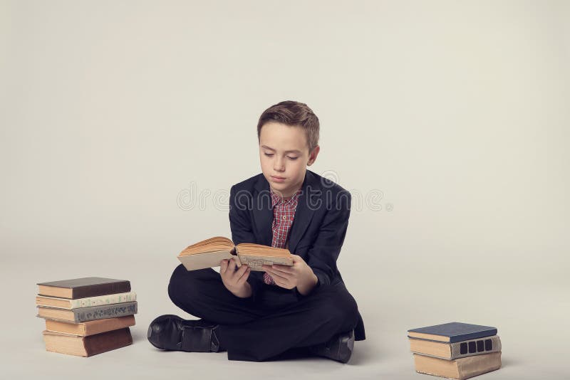Netter Junge in einer Klage, die ein Buch auf einem grauen Hintergrund sitzt und hält stockfoto