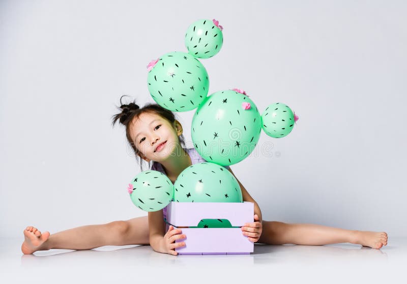 Cheerful asian girl sits takes the splits behind the box with cactus balloon with pink flowers in it, peeks out and smiles nice. Creative idea minimal concept on gray background. Cheerful asian girl sits takes the splits behind the box with cactus balloon with pink flowers in it, peeks out and smiles nice. Creative idea minimal concept on gray background