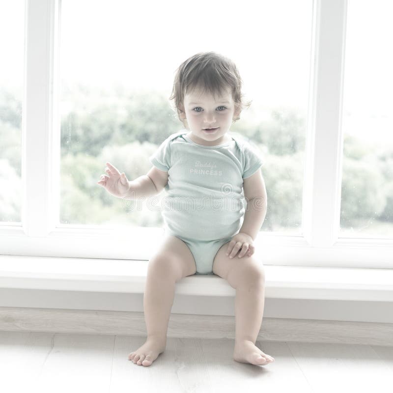 Nettes Baby zu Hause im Reinraum sitzt nahe Fenster Das schöne Baby könnte ein Junge oder ein Mädchen sein und trägt Körperanzug stockbilder