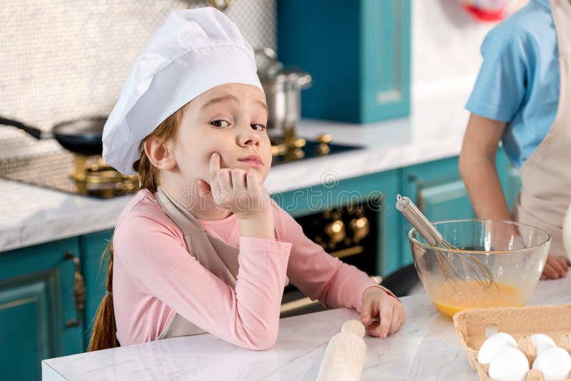 nettes Kind im Chefhut und Schutzblech, das mit der Hand auf Kinn sitzt und Kamera betrachtet stockfotos nettes Kind im Chefhut und Schutzblech, das mit der Hand auf Kinn sitzt und Kamera betrachtet stockfotos