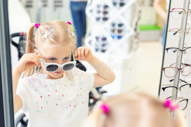 Cute little young caucasian blond girl trying on and choosing sunglasses in front of mirror at optic eyewear store. Adorable schoolgirl child having fun bying sun uv protection glasses at shop, kid, sale, happy, female, person, childhood, people, customer, lifestyle, optician, eyeglass, eyesight, children, beautiful, spectacle, care, frame, retail, design, smiling, optical, buy, eyecare, vision, portrait, shopper, cheerful, fashion, fit, choose. Cute little young caucasian blond girl trying on and choosing sunglasses in front of mirror at optic eyewear store. Adorable schoolgirl child having fun bying sun uv protection glasses at shop, kid, sale, happy, female, person, childhood, people, customer, lifestyle, optician, eyeglass, eyesight, children, beautiful, spectacle, care, frame, retail, design, smiling, optical, buy, eyecare, vision, portrait, shopper, cheerful, fashion, fit, choose