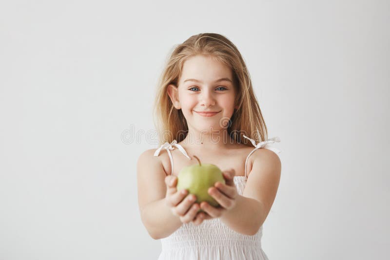 Nettes kleines Mädchen mit dem blonden langen Haar und den blauen Augen im weißen Kleid brightfully lächelnd, Apfel in den Händen