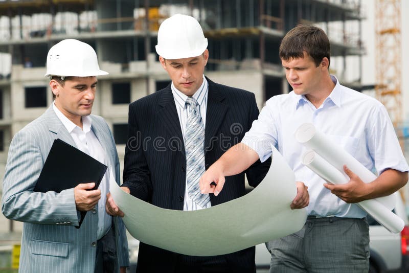 New project. Portrait of three builders standing at building site and discussing new project held by one of men New project. Portrait of three builders standing at building site and discussing new project held by one of men