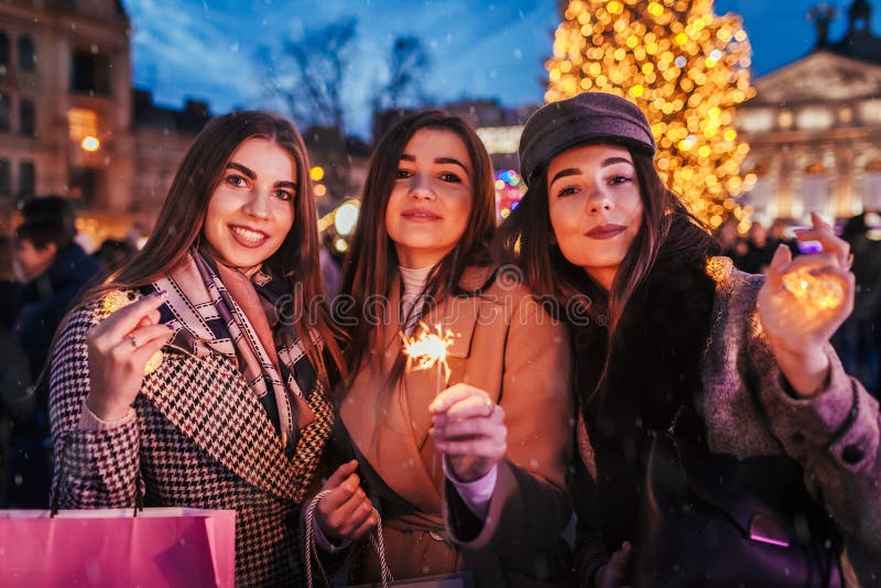 New Year concept. Women friends burning sparklers in Lviv by Christmas tree on street fair. Girls holding shopping bags stock image New Year concept. Women friends burning sparklers in Lviv by Christmas tree on street fair. Girls holding shopping bags stock image