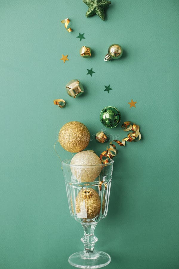 New year party concept. Stylish christmas gold baubles and confetti pouring from champagne glass on green background flat lay.