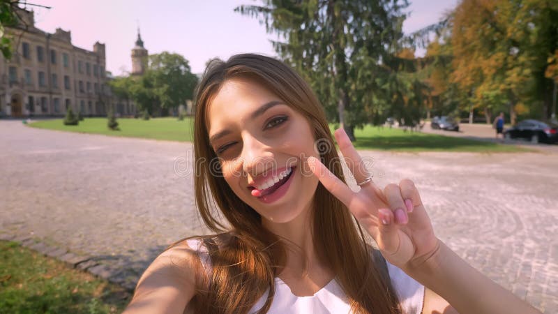 Nice de jonge vrouw zich in park in dag bevindt, houdt camera, maakt selfie, toont vredesteken, communicatie concept