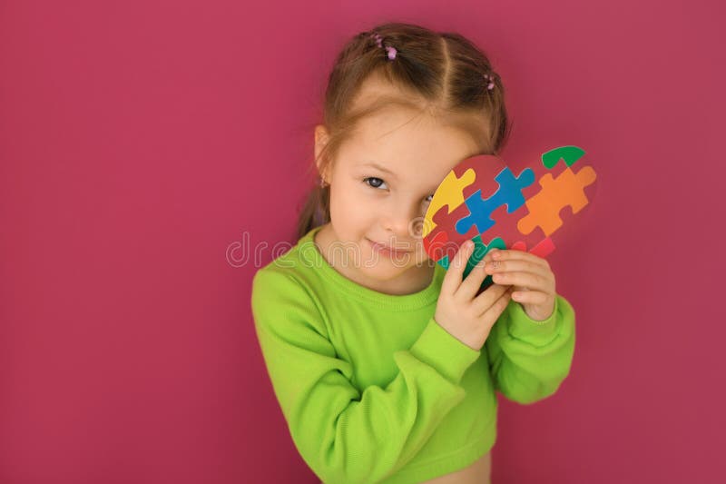 Nice girl looks into camera smiling and holding heart card with colored puzzles